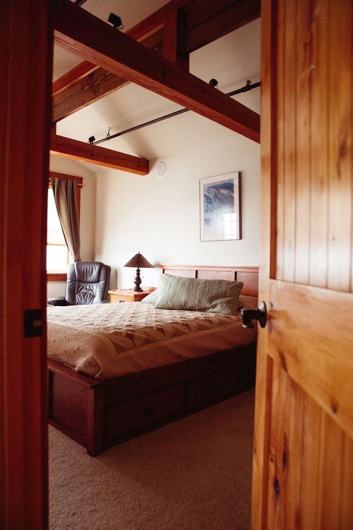 Bedroom in The Loft seen through an open timber door, with a queen bed, reading lamp and recliner