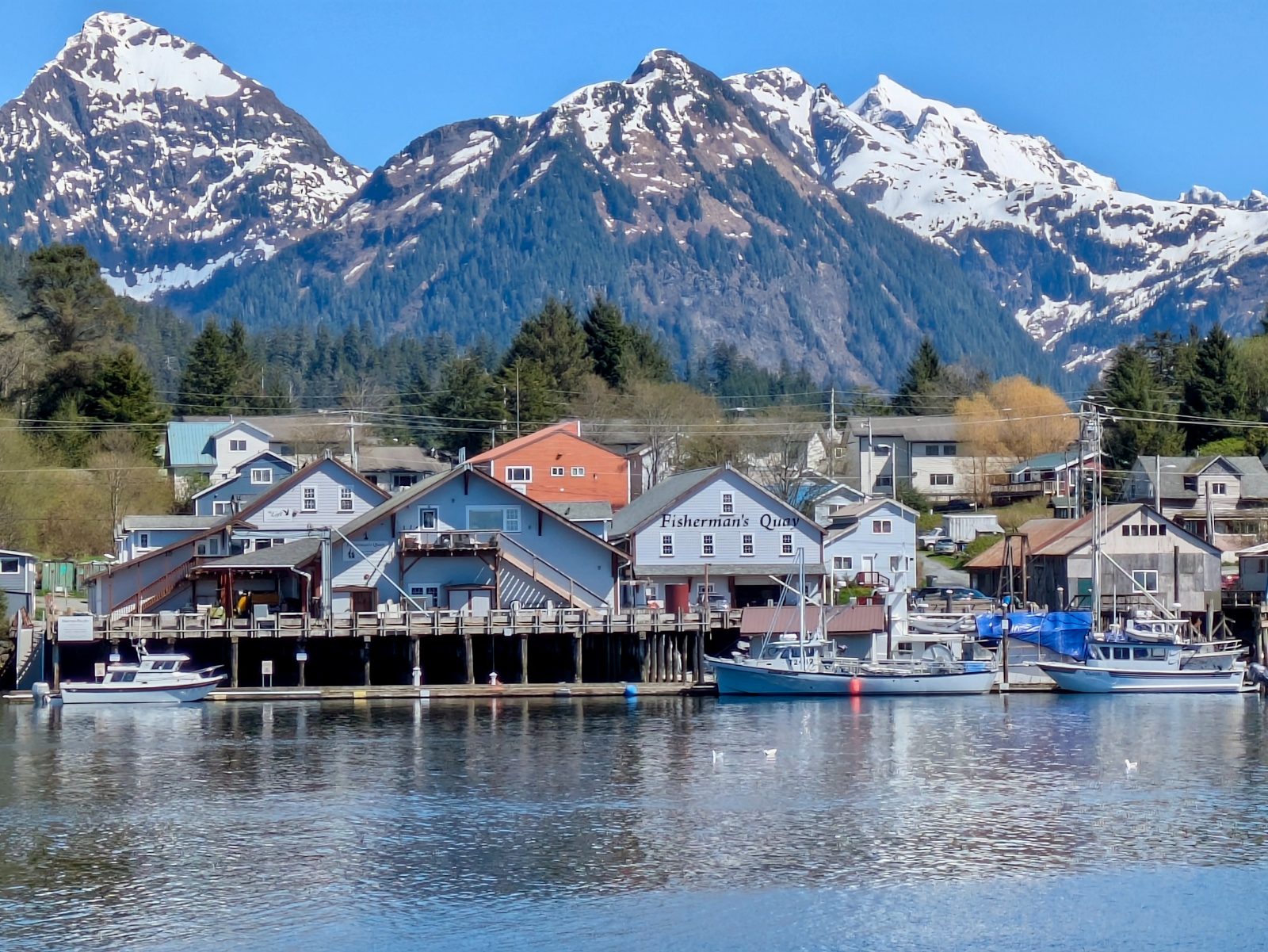 The Fisherman's Quay buildings on the Sitka waterfront seen from across the channel, with fishing boats along the dock and snow-covered mountains behind