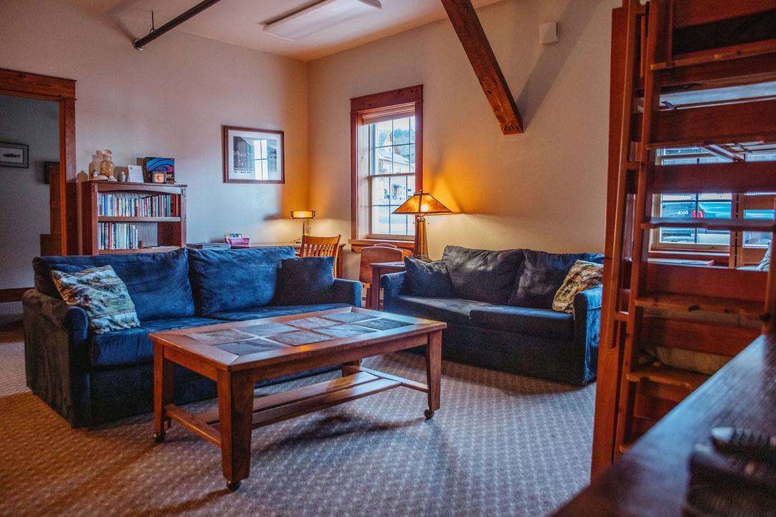Main room of the South Bunkhouse with two blue sofas facing a wooden coffee table, a bookshelf and lamps, and a timber bunk bed at the right